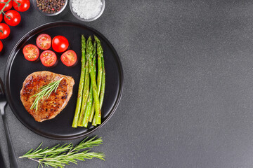 Tasty grilled beef steak served with green asparagus and cherry tomatoes on black plate on gray textured backgound. Top view, copy space. Perfect for food, diet, or culinary design concepts.