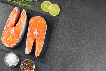 Two fresh raw salmon steaks on black stone surface with food ingredients. Tomatoes, lime, rosemary,...