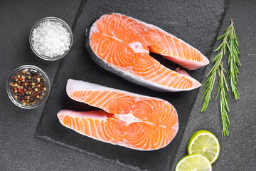 Two fresh raw salmon steaks on black stone surface with food ingredients. Tomatoes, lime, rosemary,...