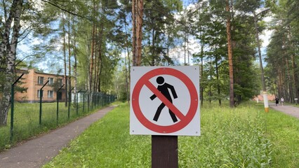 Close-up of no pedestrian sign on forest path with fence and buildings in background, sunny day,...