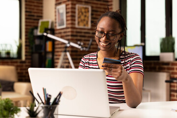 Young female adult seated indoors using credit card to complete digital payment on laptop. Smiling...