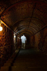 A man descends a staircase through a dark stone tunnel lit by lamps