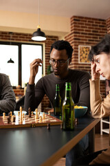 African american man sitting with friends, playing chess and snacking in brick wall room. Pensive black male individual looking at chessboard, planning next move during intense friendly board game.