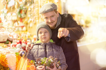 Man and son walk through New Year fair, stare at attractive goods, souvenirs. They look into...