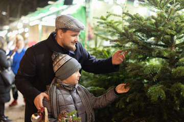Father and son choose a lush Christmas tree for home. Joyful man with son near market with fir trees