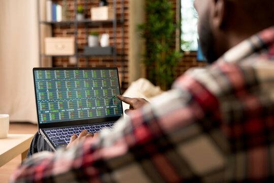 Remote trader sitting in living room, tracking stock indexes and cryptocurrency data on laptop. African american man working from home, reviewing trading market trends and investment strategies.