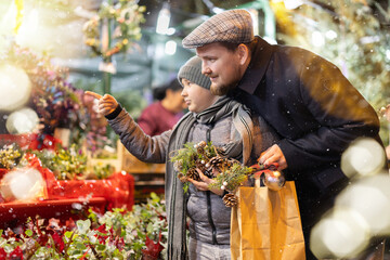 Father and son stand near the counter at the Christmas market, choosing accessories, flower...