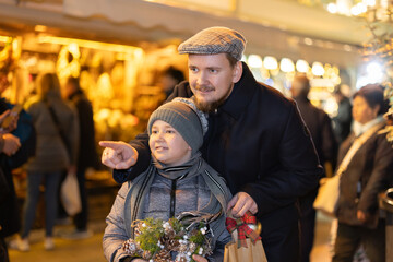 Boy with father stands at fair decorated for New Year holidays, examine surrounding festive...