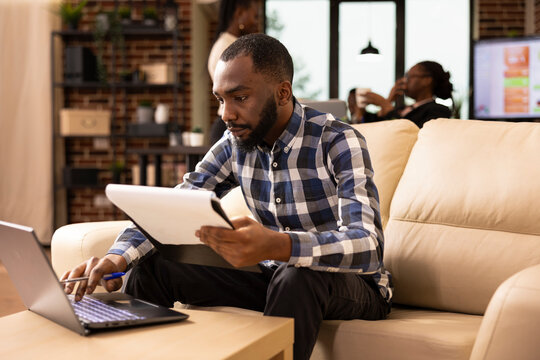 Portrait of black businessman reviewing financial documents while checking related details on his laptop. African american male professional holding clipboard, engaged in startup project task. - Powered by Adobe