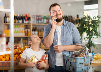 Smiling young bearded man holding basket with groceries and talking on phone during casual shopping trip with cheerful preteen son, clutching snacks and juice, in local supermarket