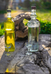 Two bottles of liquor, one yellow and one transparent, next to two small glasses on a rustic wooden table in the open air, with vegetation in the background