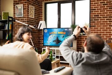 White man and woman relax together, celebrating shared video game victory. Young caucsasian adults with raised hands, cheering and enjoying drinks on sofa while TV flashes winning message.