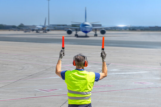Aircraft marshaller in PPE guiding airplane on runway with marshalling wands at airport.