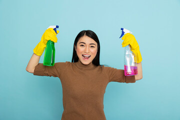 Energetic asian housekeeper in rubber gloves holding colorful cleaning sprays and flexing arms,...
