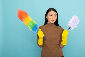 Confident female housekeeper holding dusting tools stands ready for cleanup tasks, isolated on blue background. Young adult woman represents modern housekeeping and cleanliness standards.
