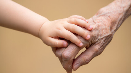 Fototapeta premium Extreme close-up of a young child's smooth hand gently holding the wrinkled hand of an elderly person against a warm, neutral beige background. 