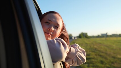 Dreaming young woman leans out of car window travelling across countryside at sunset closeup....