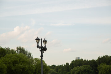 Street black iron design lamp in the park outdoors on summer green trees background under blue day sky with clouds