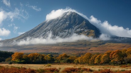 Fototapeta premium Mountain peak veiled in clouds, autumn trees below, blue sky backdrop