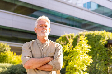Mature businessman standing outdoors with arms crossed in front of office building. Confident expression in bright daylight with greenery around