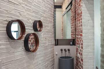 Industrial-style bathroom corner with exposed red and white brick walls, round metal-framed...