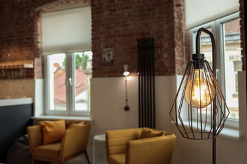 Close-up of an industrial-style floor lamp with a glowing Edison bulb in a metal cage frame, set in...