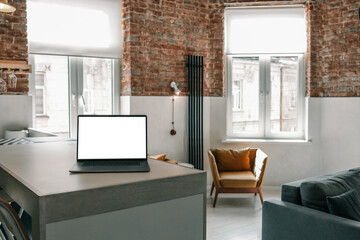 Modern loft-style living room with a laptop on the kitchen counter, exposed brick walls, cozy armchair, and natural light coming through large windows