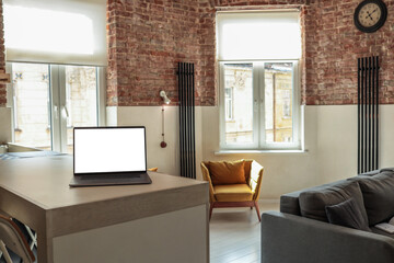 Modern loft-style living room with a laptop on the kitchen counter, exposed brick walls, cozy armchair, and natural light coming through large windows