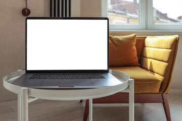 Close-up of a blank screen laptop on a small round table in a modern cozy interior with sunlight, mustard armchair, and large window in the background