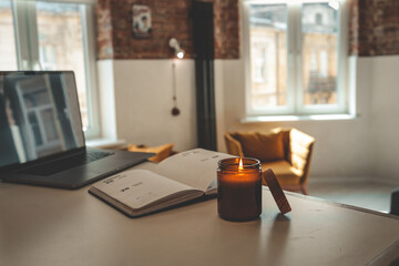 Burning candle and open planner on a table in cozy loft interior with warm light, mustard chairs,...