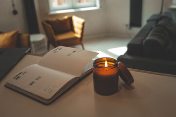 Burning candle and open planner on a table in cozy loft interior with warm light, mustard chairs,...