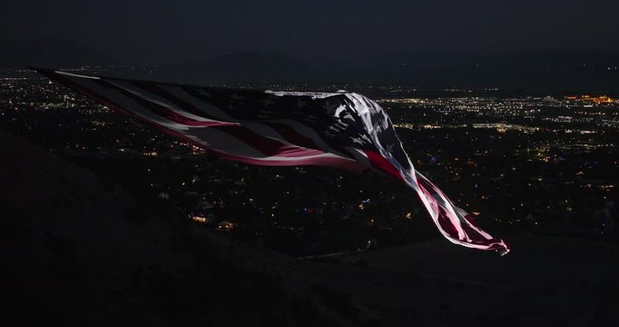 Drone shot of rippling American flag with spotlight as it flys over valley with scattered fireworks at night