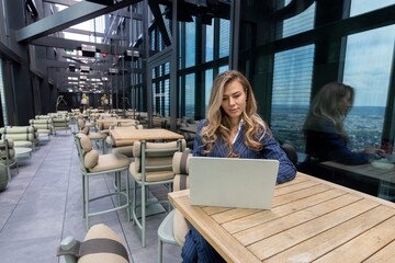 A young woman is intently focused on her laptop in a stylish rooftop cafe that boasts breathtaking...