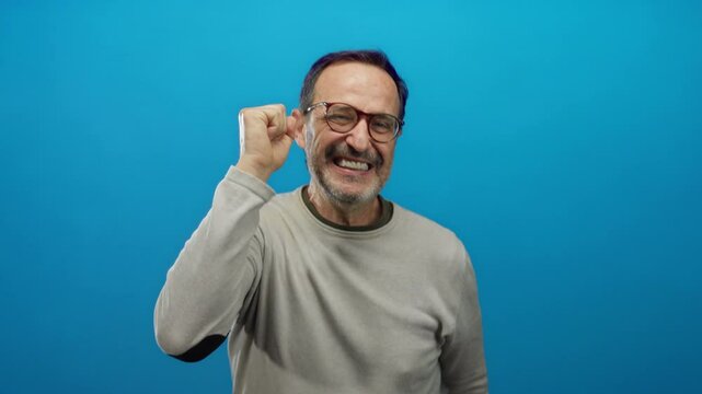 Mature hispanic man expressing joy and excitement with raised hands against a vibrant blue background, showcasing an enthusiastic and animated expression.