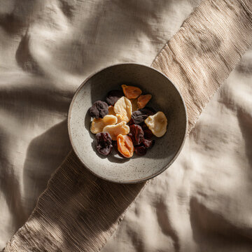 Dried fruits above view, ceramic bowl with dried fruit pieces on natural linen runner, minimalist food style, warm neutral palette