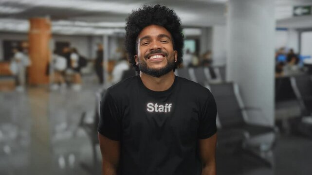 Man in black staff uniform smiling indoors at a busy airport, embodying friendliness and professionalism in a diverse, vibrant setting. - Powered by Adobe
