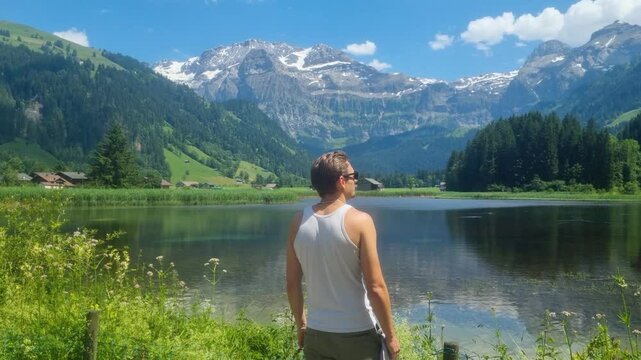 A muscular man in a white tank top gazes out over a tranquil lake toward the majestic Wildstrubel and surrounding Alpine peaks on a sunny day in Lenk, Switzerland
