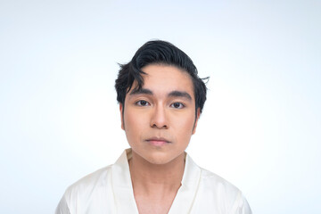 Young Southeast Asian man in a white silk robe stares directly at the camera with a focused,...