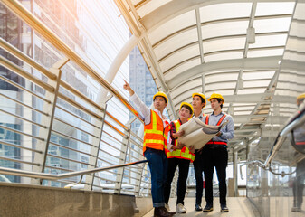 Four Asian construction engineers in yellow hard hats and high visibility safety vests examine blueprints on modern walkway. Team leader points upward while discussing project planning details 