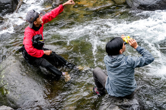 Portrait Of Two Adventures Sitting On Rocky River While Looking View In Nature With Binoculars