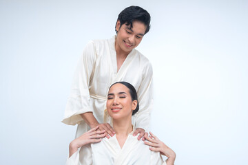 Young Asian man gives massage to woman in silk robe. She closes eyes in calm delight while he looks...