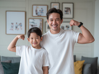 young boy and his father flex their muscles, both smiling joyfully in matching white t shirts. cozy living room features family photos in background, creating warm atmosphere