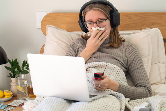 Remote work while sick. Woman in bed sneezing with laptop and headphones. Health care
