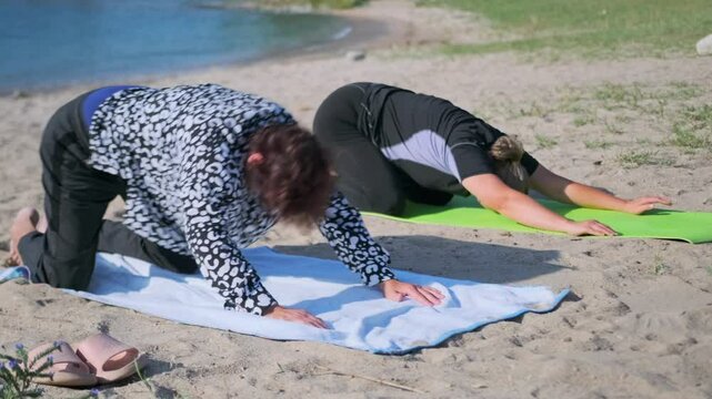 Two women doing yoga stretch on a sandy beach, lying on bellies and lifting their upper bodies supported by their hands. This pose opens the chest and stretches the back beside a natural waterside.