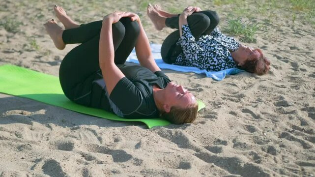 A mature mother and her adult daughter lie on mats at a sandy beach, gently hugging their knees to the chest in a calming yoga stretch that relieves tension and promotes flexibility.