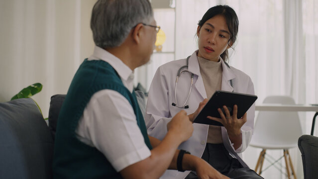 A young female doctor in a lab coat and stethoscope uses a tablet to discuss medical records with an elderly male patient in a bright clinic.
