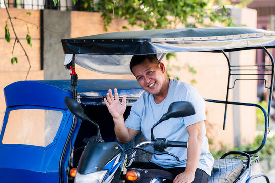 A cheerful tricycle driver sits on his vehicle and waves at the camera with a big, wide smile in a friendly gesture.