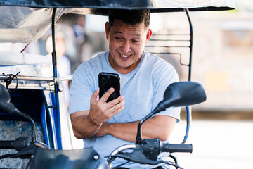 A smiling Southeast Asian tricycle driver sits in his vehicle while chatting on a video call,...