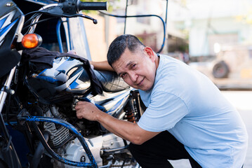 A middle-aged Southeast Asian tricycle driver kneels down fixing his motorcycle while giving a...