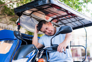 Obraz premium Asian man wearing a blue shirt looks irritated and disappointed while scratching his head, clearly frustrated with his tricycle’s motor not starting.